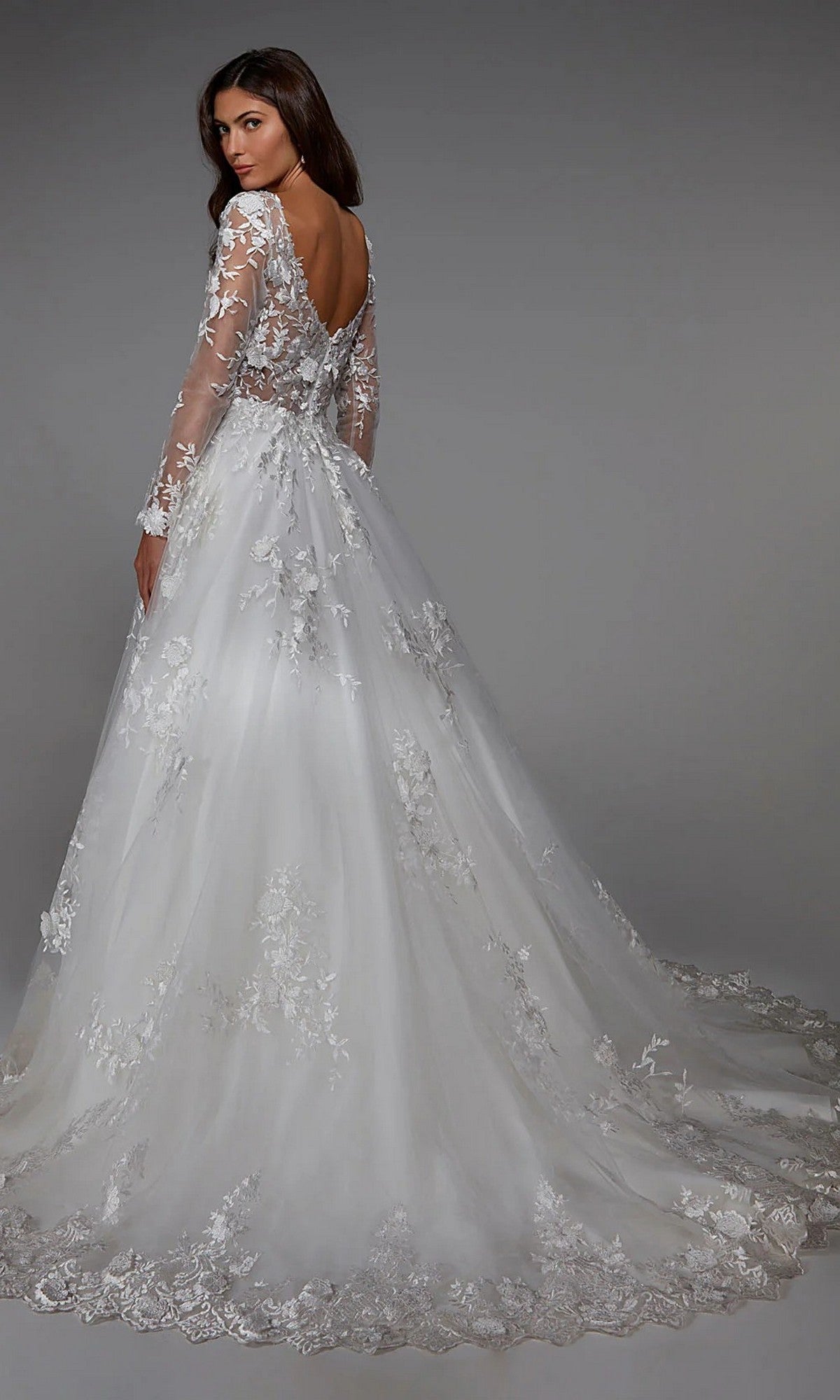 A woman models the Alyce 7046 long white dress, featuring lace, floral embroidery, long sleeves, a deep V-back, and a flowing train, standing against a plain gray background.